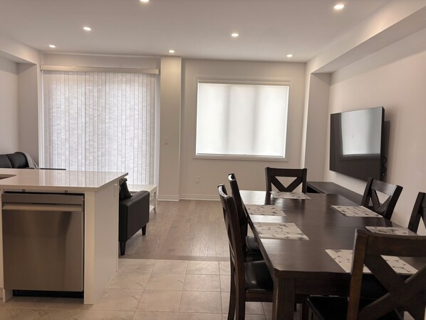 A modern hotel room with a dining area, a flat-screen TV, and a large window with blinds.