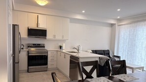 Une cuisine moderne avec des meubles blancs, des appareils électroménagers en acier inoxydable, un coin repas avec une table et des chaises en bois foncé, et une fenêtre avec des stores.