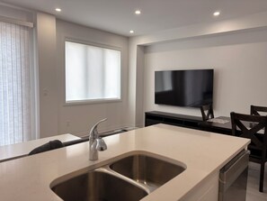 A modern kitchen with a double sink, a dining area with a TV, and a window with blinds.