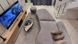 A modern living room with a curved grey sofa, a flat-screen TV, a decorative table, and a staircase. - Poise And Class Apartment (Lagos)