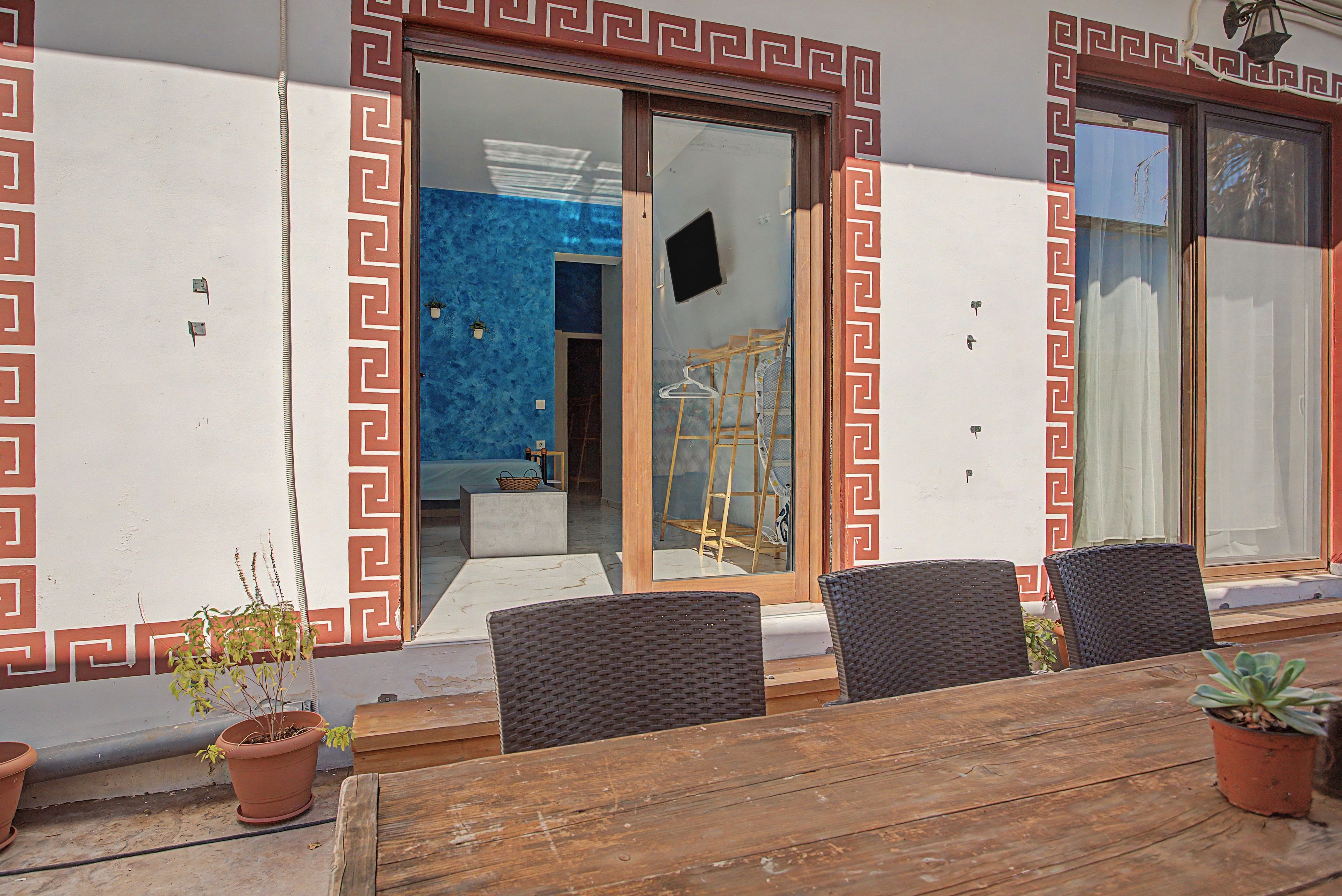 An outdoor seating area with a wooden table and chairs, potted plants, and a blue interior visible through a glass door.