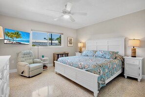 A bedroom with a bed, a recliner, a ceiling fan, a window with a view, and a nightstand with a lamp. - Jamaica Royale 106 Beautiful Condo! (Siesta Key)
