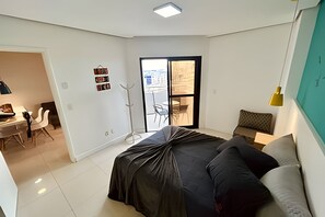 A modern bedroom with a bed, a desk, and a chair. There is a balcony with a view of the city. - SA Clube XV  Apto CXV1601 (Santos)