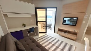 Una sala moderna con un sofá, una televisión, un escritorio pequeño y un ventanal corredero que da a un balcón.