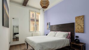 Ein Schlafzimmer mit einem Bett, einem Nachttisch, einem Spiegel und einem Fenster mit Blick auf Gebäude.