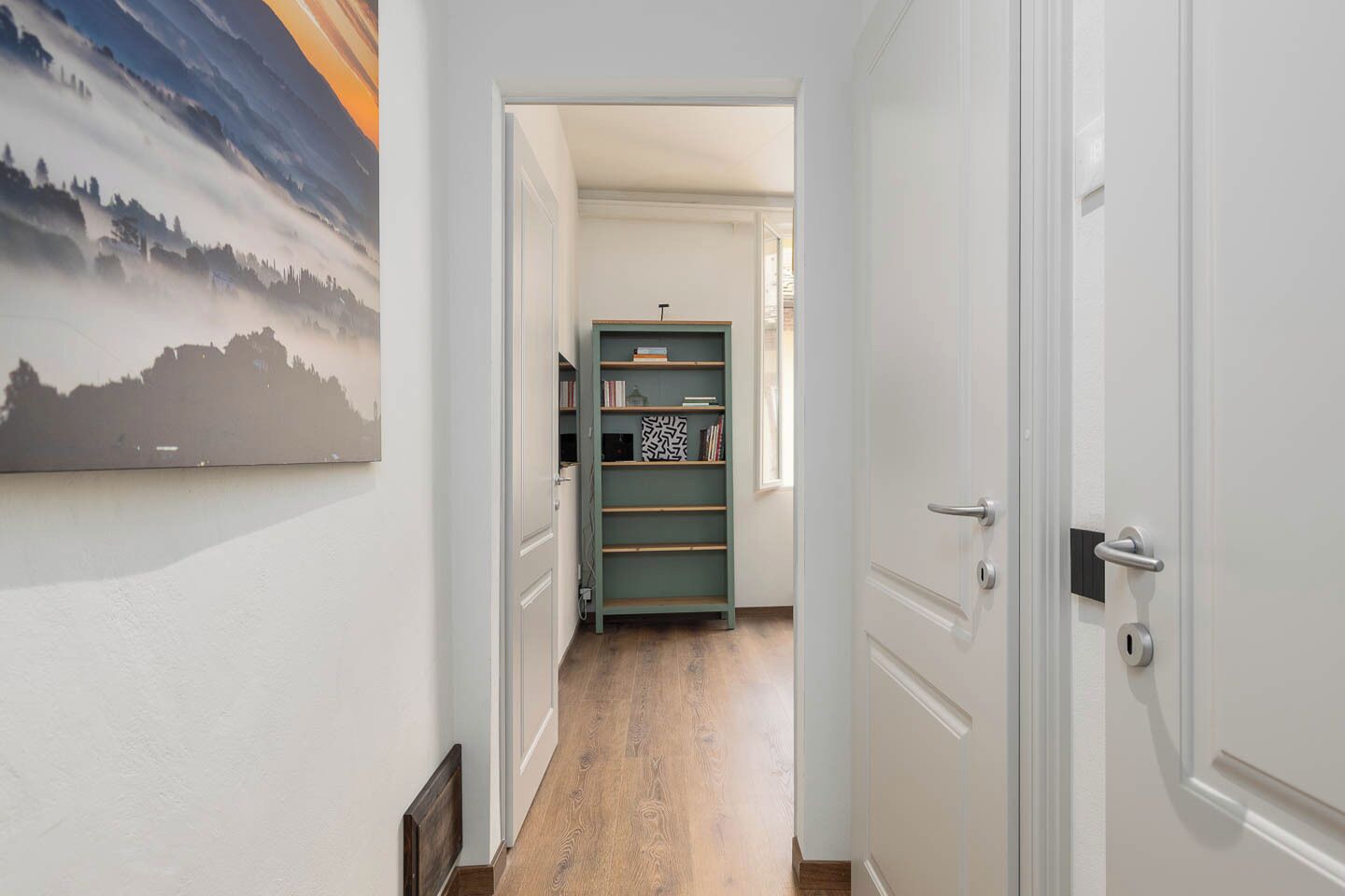Un pasillo con piso de madera, una estantería verde y una puerta con picaporte moderno.