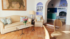Een woonkamer met een bank, salontafel, fauteuils, een boekenkast en een keuken die zichtbaar is via een boogvormige doorgang.