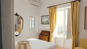 Een slaapkamer met een bed, een lamp, een dressoir, een stoel en een raam met gordijnen.