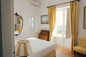A bedroom with a bed, a lamp, a chest of drawers, a chair, and a window with curtains. - Villa Angie by My Villa Collection (Positano)