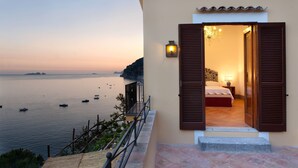 Un balcone con vista sul mare e sulle barche, una camera da letto con un letto e un comodino, e una lampada da parete accesa.