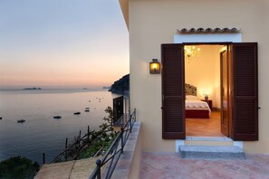 Ein Balkon mit Meer- und Bootsblick, ein Schlafzimmer mit Bett und Nachttisch sowie eine beleuchtete Wandlampe.