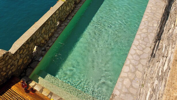 Una piscina rettangolare con pareti in pietra, un solarium in legno e lettini da relax.
