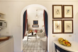 Ein Wohnzimmer mit einer Couch, einem Couchtisch, einer Obstschale, gerahmten Bildern an der Wand und einer Mikrowelle in der Küchenzeile.