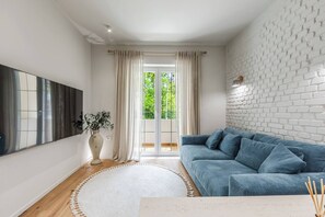 A modern living room with a blue sofa, a large window with curtains, a white brick wall, and a flat-screen TV mounted on the wall. - The Homey Flat | Stylish 2BR City Apartment by PikHost (Tiranë)