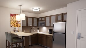A modern kitchen with wooden cabinets, a stainless steel refrigerator, a microwave, and a sink. There is a dining table with a chair and a pendant light.