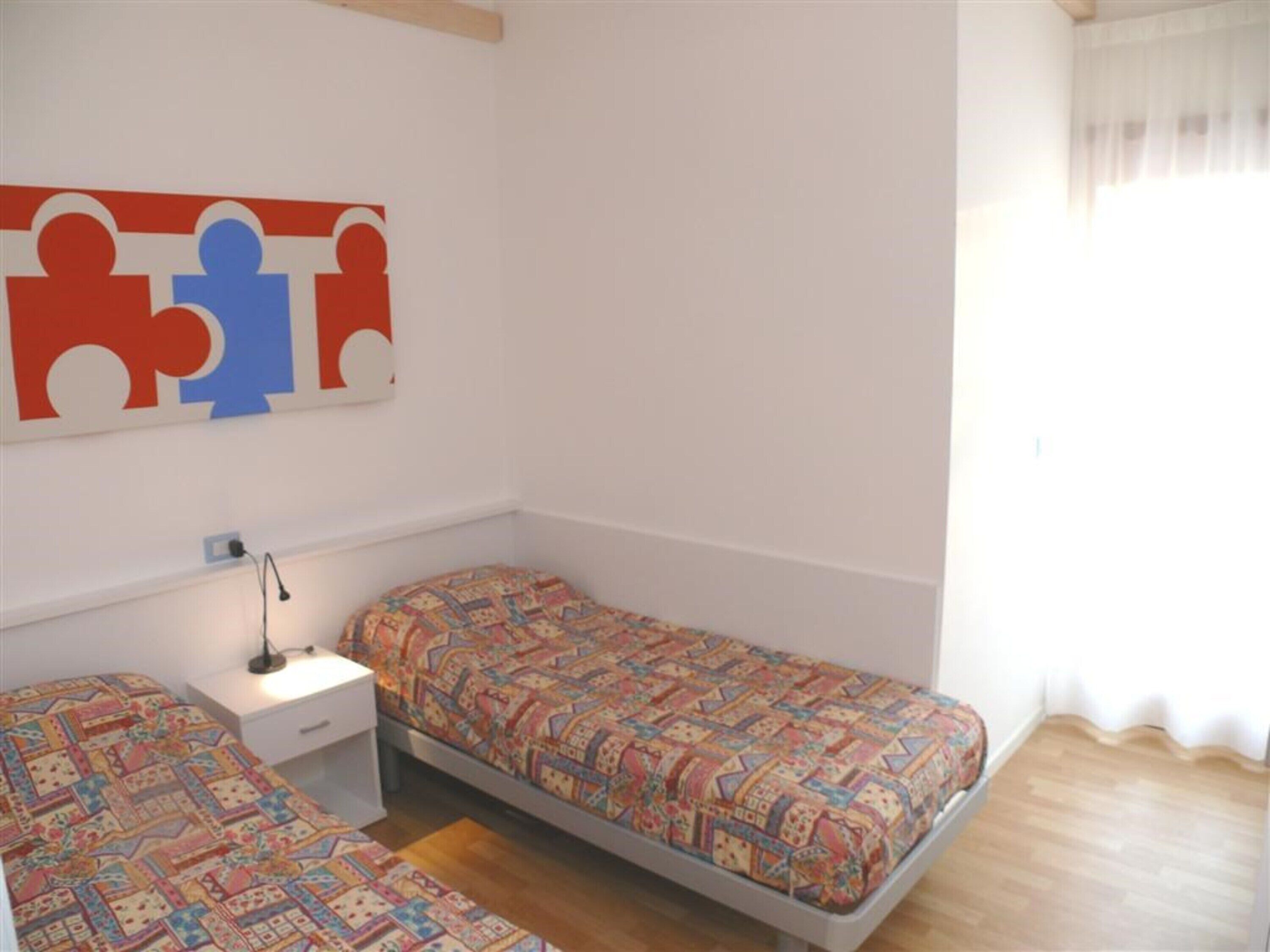 Quarto com duas camas de solteiro, uma mesinha de cabeceira com abajur, uma obra de arte na parede e uma janela com cortinas.