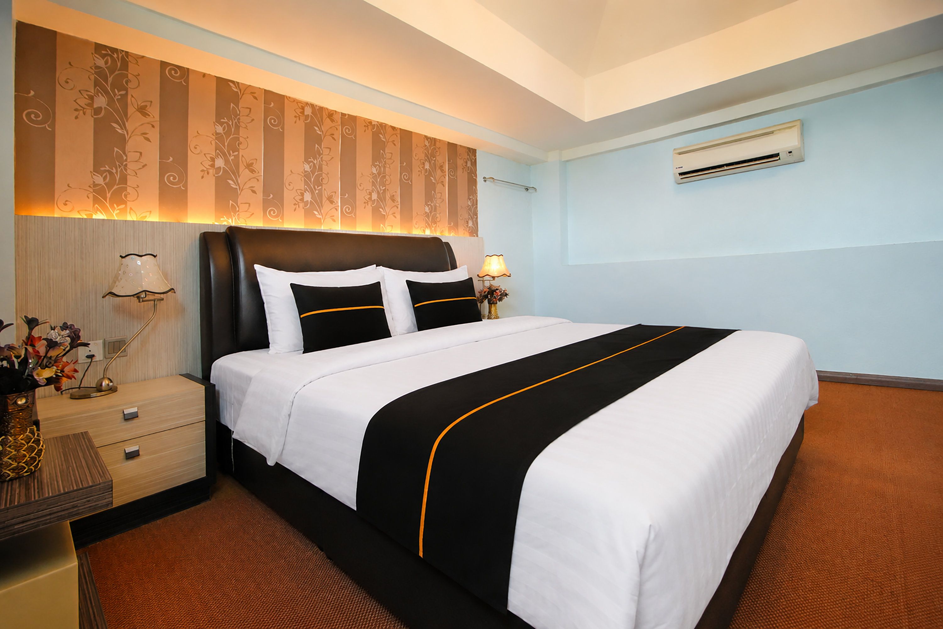 A hotel room with a large bed, a nightstand with a lamp, and a wall-mounted air conditioner.