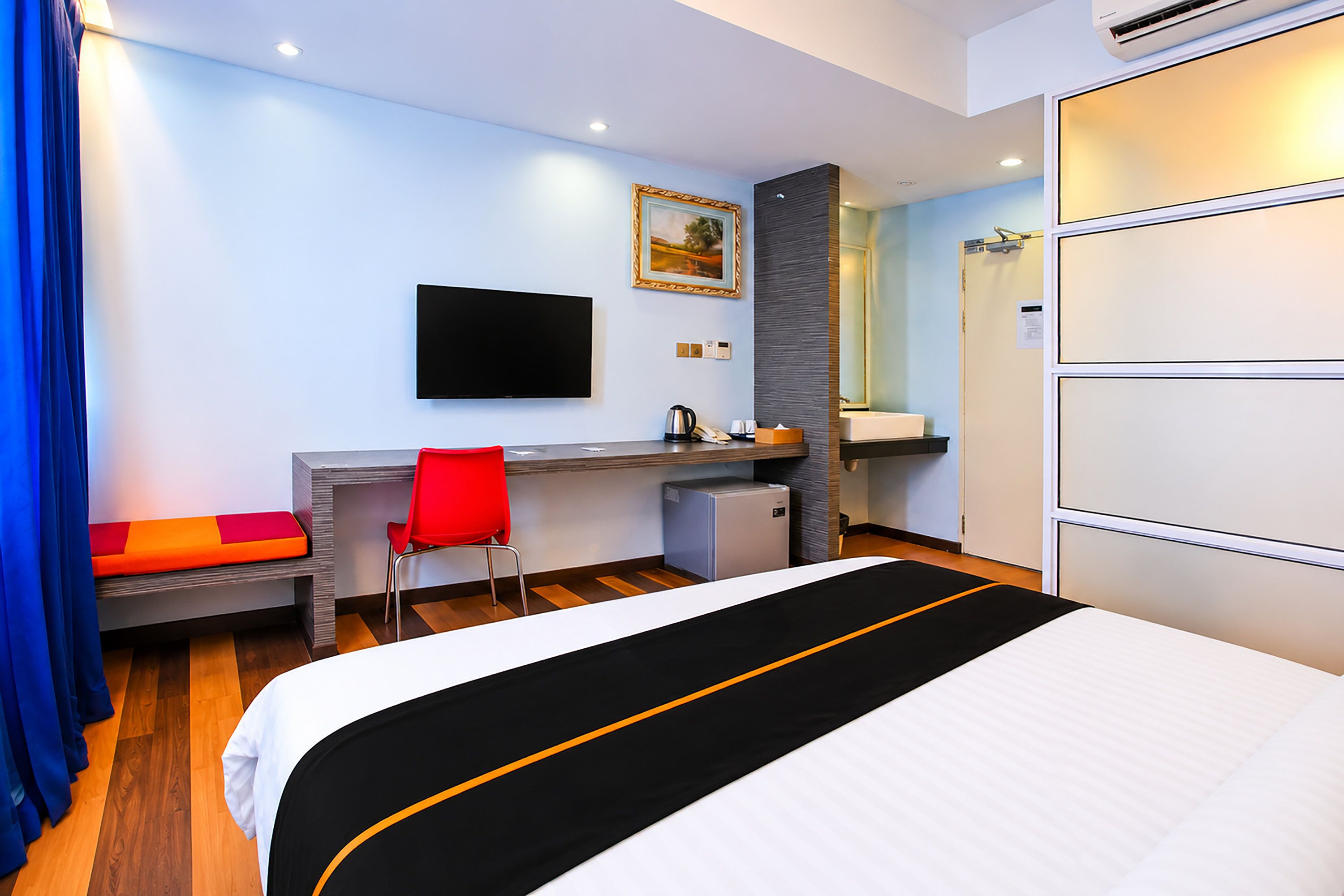 A hotel room with a bed, a desk with a chair, a television, a small refrigerator, and a bathroom with a mirror.
