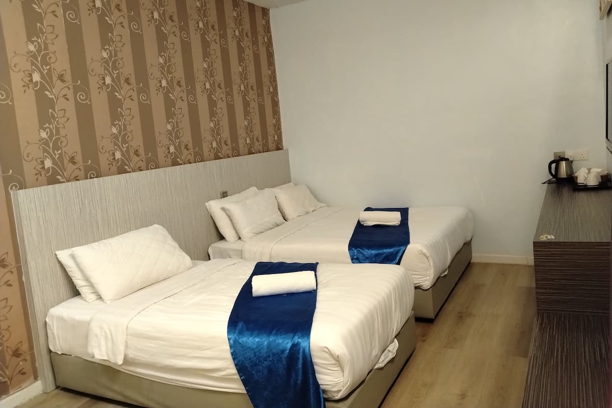 A hotel room with two beds, a wooden headboard, a blue throw blanket, a wall with floral wallpaper, and a small table with a kettle and cups.