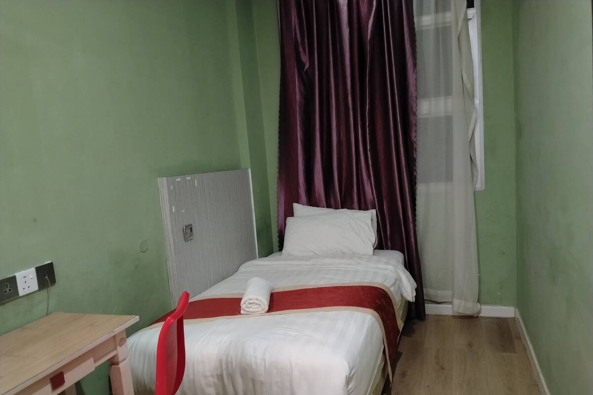 A small, single-bed room with a wooden desk, a red chair, and a window with purple curtains.