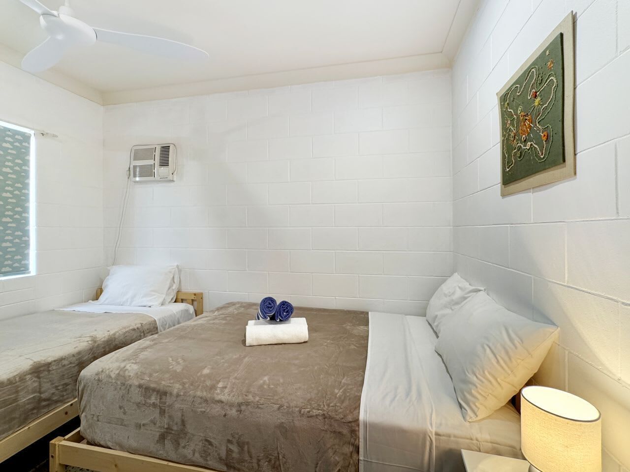 Una habitación con dos camas, ventilador de techo, aire acondicionado y una obra de arte en la pared.
