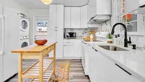 Cozinha moderna com armários brancos, mesa de madeira, máquina de lavar, micro-ondas e pia com torneira preta.