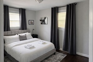 Ein modernes Schlafzimmer mit einem großen Bett, grauen Vorhängen, einem Kopfteil, einem Nachttisch und einem gerahmten Bild an der Wand.
