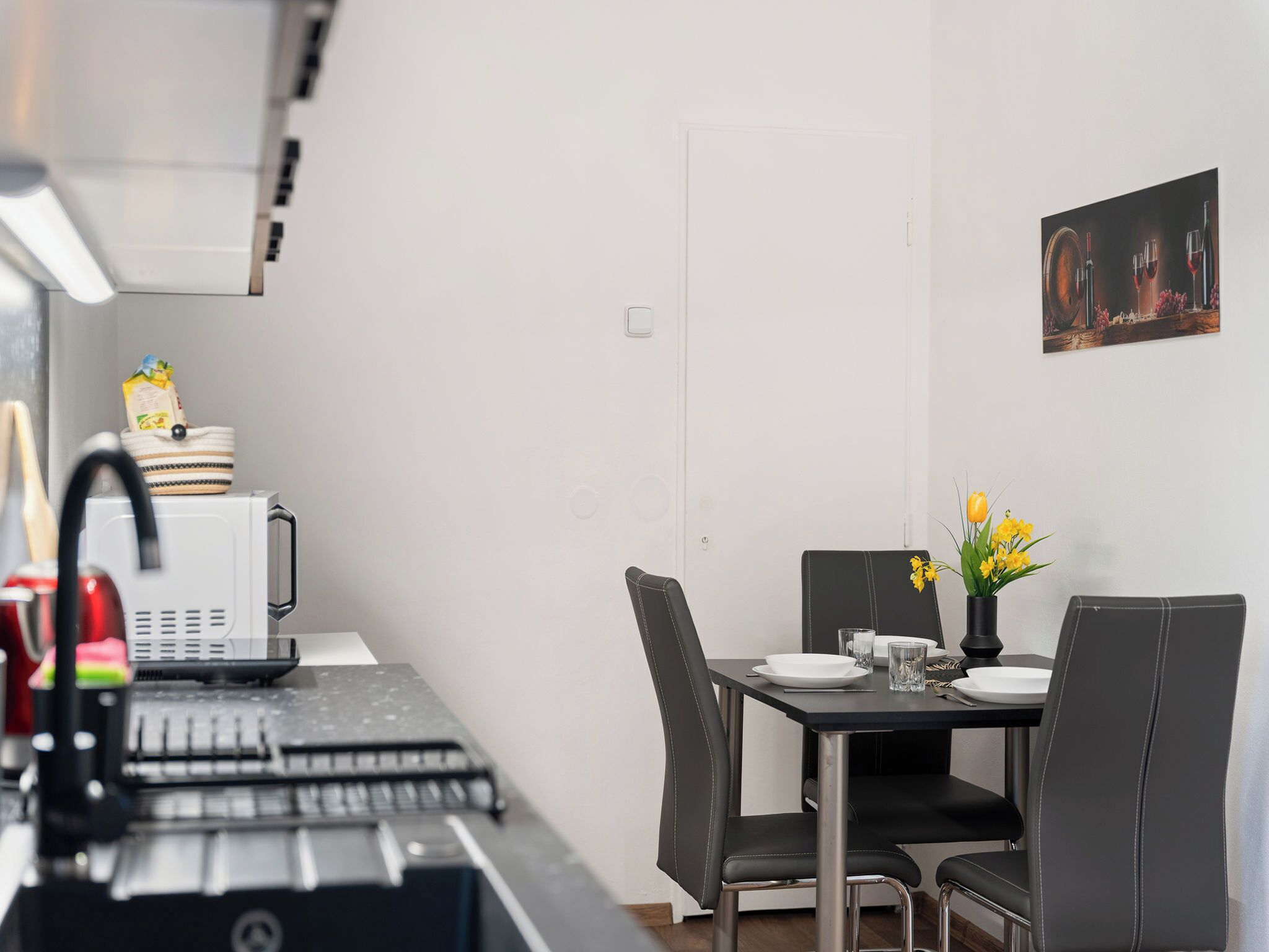 Une cuisine compacte équipée d’un four à micro-ondes, d’une bouilloire et d’une machine à café. Une table à manger, avec des chaises et un vase de fleurs, est placée contre un mur sobre.