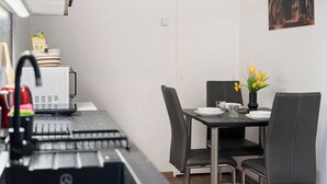 A compact kitchen with a microwave, kettle, and coffee machine. A dining table with chairs and a vase of flowers is set against a plain wall. - Happy Home by Interhome (Hradesice)