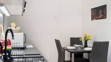 Une cuisine compacte équipée d’un four à micro-ondes, d’une bouilloire et d’une machine à café. Une table à manger, avec des chaises et un vase de fleurs, est placée contre un mur sobre.