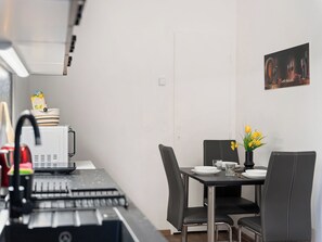 Une cuisine compacte équipée d’un four à micro-ondes, d’une bouilloire et d’une machine à café. Une table à manger, avec des chaises et un vase de fleurs, est placée contre un mur sobre.