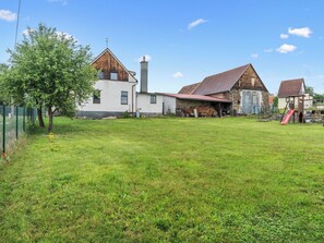 A rural property with a large grassy yard, a white building with a wooden extension, a stone barn, and a wooden playhouse with a slide.