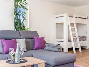 Ein Zimmer mit grauem Sofa, einem weißen Stockbett, einem Holztisch mit einer Vase und Gläsern sowie einer großen Pflanze.