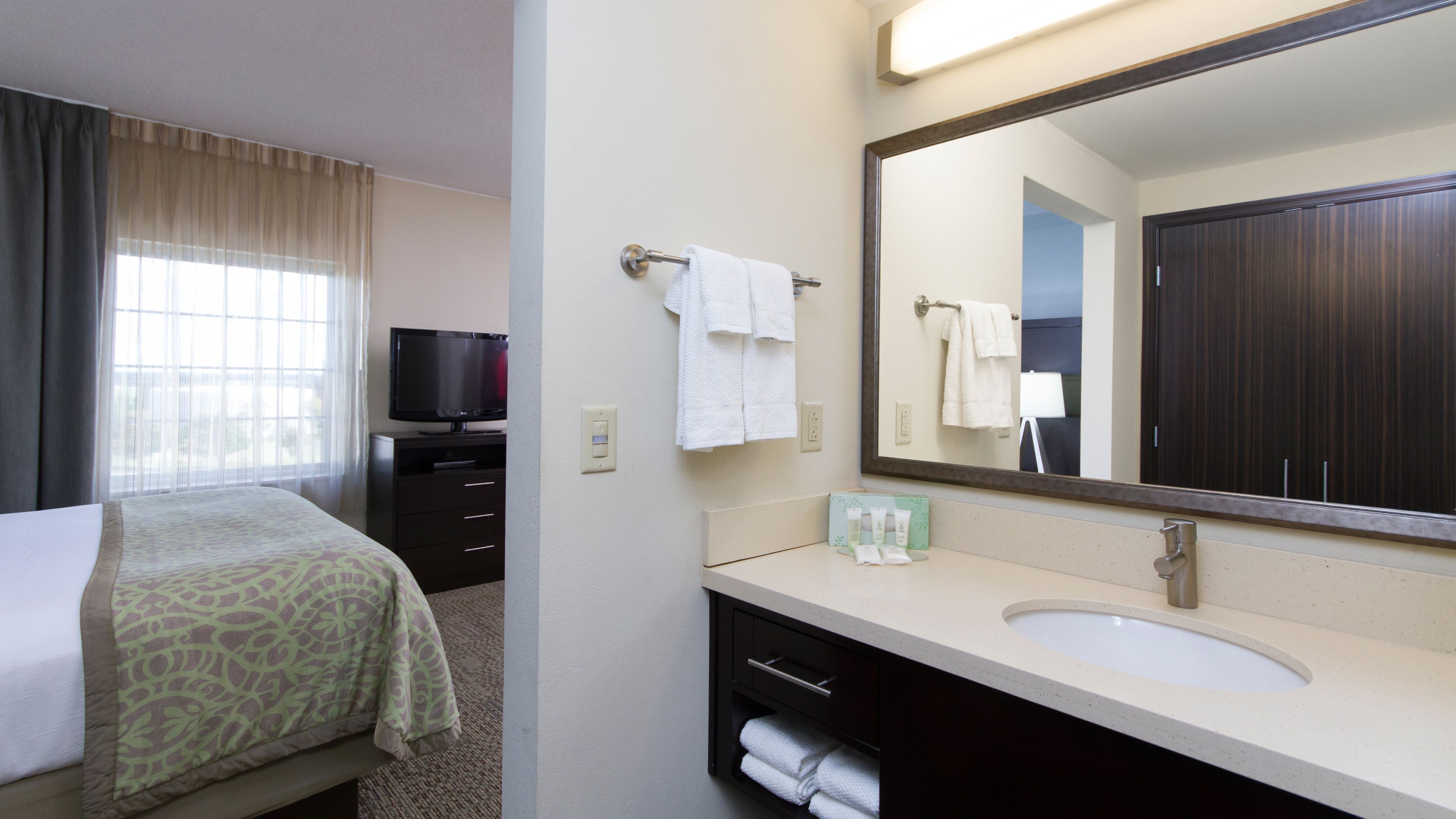 A hotel room with a bed, a TV, a large mirror, a sink, and a window with curtains.