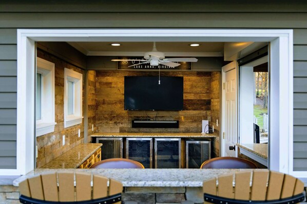 An outdoor kitchen with a stone backsplash, a flat-screen TV mounted above the counter, and a ceiling fan.
