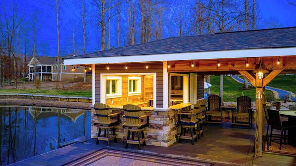 A covered outdoor patio with stone walls, wooden furniture, and a view of a pond.