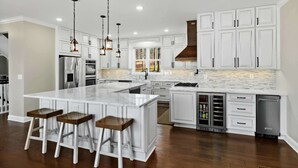 Une cuisine moderne avec des meubles blancs, un grand îlot central doté de tabourets, des appareils en acier inoxydable et un éclairage suspendu.