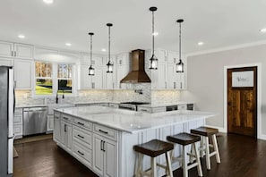A kitchen with white cabinetry, a large island with stools, a stainless steel dishwasher, and a view to the outdoors.