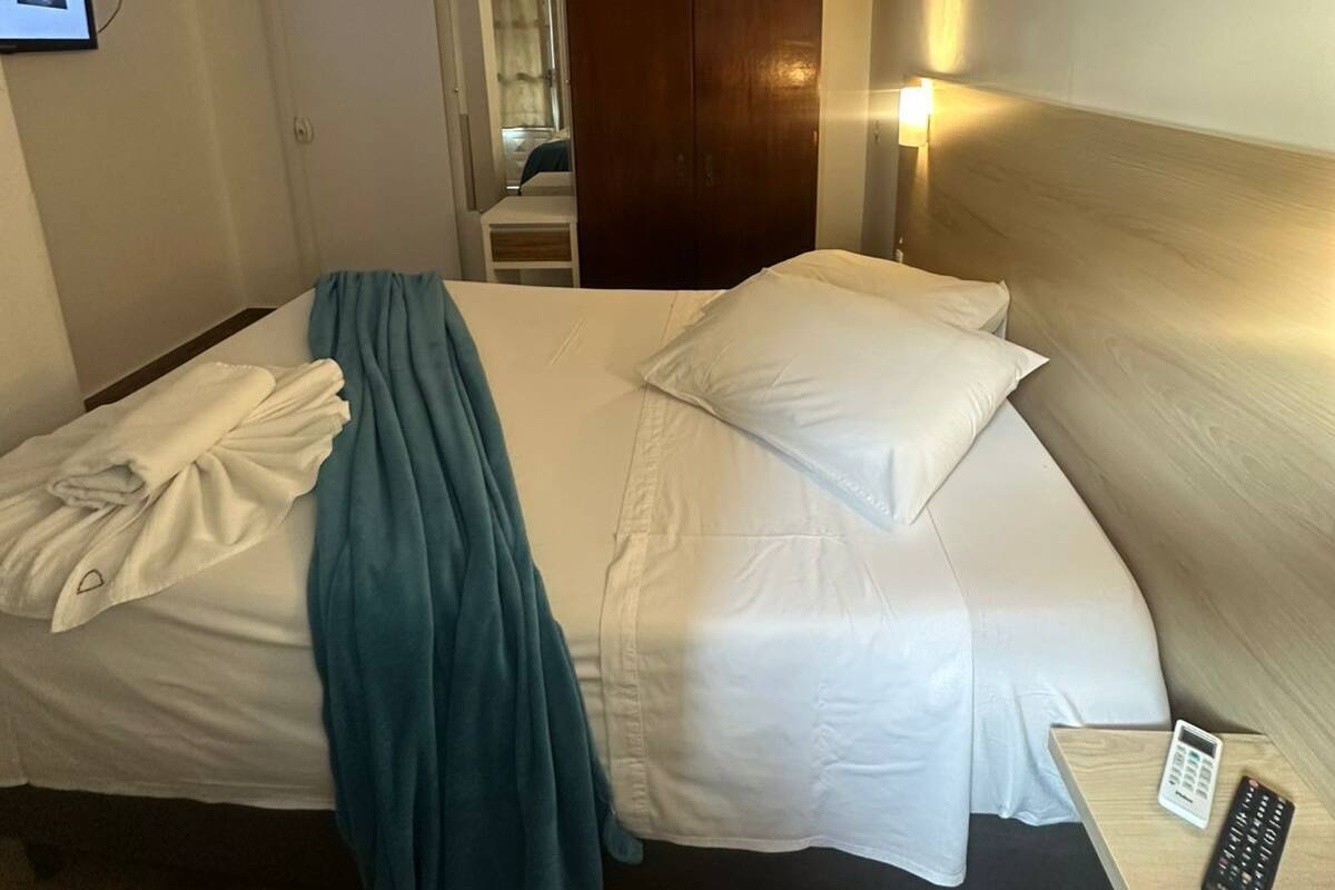 Une chambre d’hôtel comprenant un lit, une table de chevet avec une télécommande, une lampe fixée au mur et une porte donnant accès à une salle de bain.