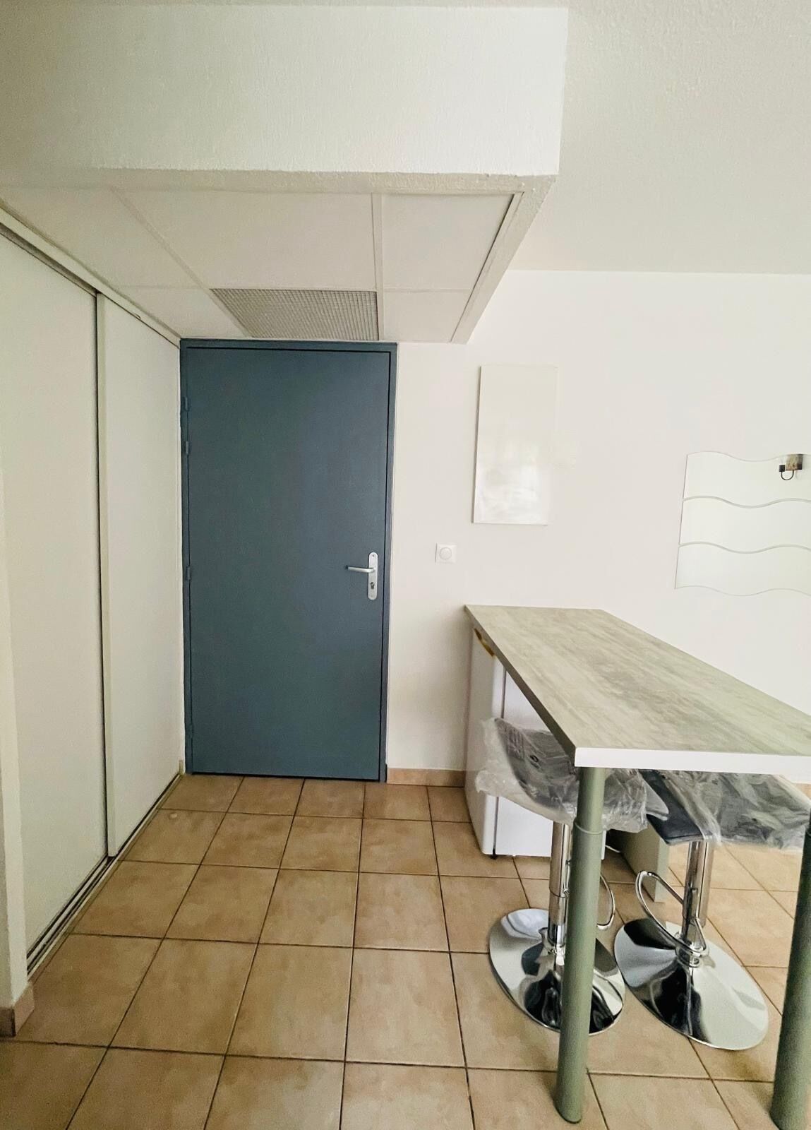 Une pièce avec une porte fermée, un sol carrelé, une table avec une chaise et un luminaire fixé au mur.