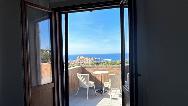 Puerta corredera abierta que da a un balcón con mesa y sillas, cama con colcha estampada y vistas al mar y edificios a lo lejos.