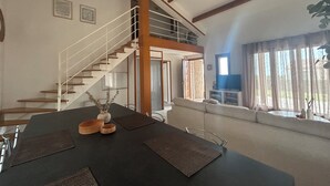 Un comedor con techo alto, vigas de madera, una escalera y un televisor sobre un mueble.