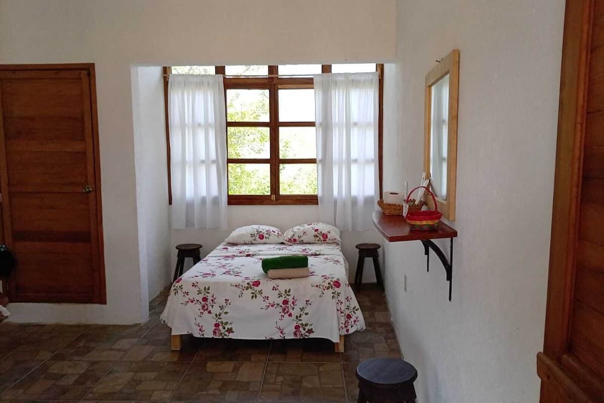 A bedroom with a bed, a window with curtains, a wooden door, a small table with a basket, and a stool.