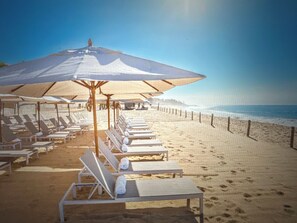 Ein Strand mit Reihen von Liegestühlen und Sonnenschirmen, feiner Sand und das Meer im Hintergrund.