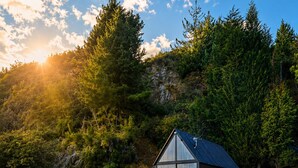 A small, dark-colored cabin with a metal roof, surrounded by lush greenery and trees, with the sun setting in the background. - Basecamp A Boutique Cabin Experience in the Wild (Carmen de Carupa)