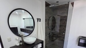 Un baño moderno con un espejo redondo, un lavabo blanco, un mueble de tocador negro, un box de ducha de vidrio y una alfombra a rayas.