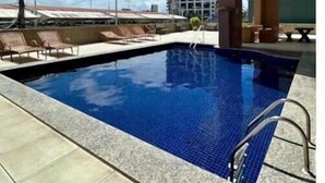 A swimming pool with tiled edges, a clear blue sky, and a modern building in the background. - 504 ocean tower 60m2 sea view 2 quartos (Fortaleza)