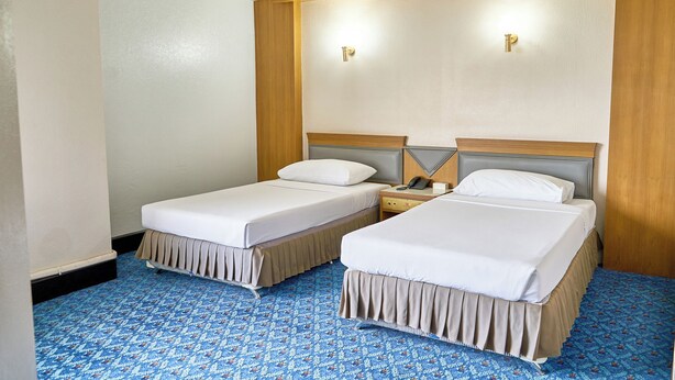 A hotel room with two single beds, a wooden headboard, a bedside table with a lamp, and a patterned carpet.