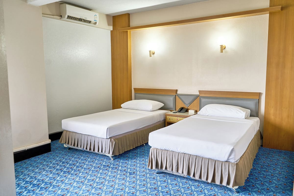 Una habitación de hotel con dos camas individuales, cabecero de madera, mesita de noche con lámpara y alfombra estampada.