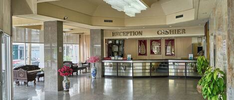 A hotel reception area with a reception desk, a cash register, and a sign that reads 'RECEPTION CASHIER.' There are potted plants and a vase with flowers on the reception desk.